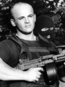 Black-and-white portrait with a machine gun; a masked man behind